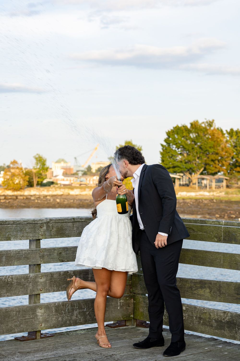 Newlywed couple kissing while popping champagne on a waterfront dock, bride in white dress, groom in black suit.