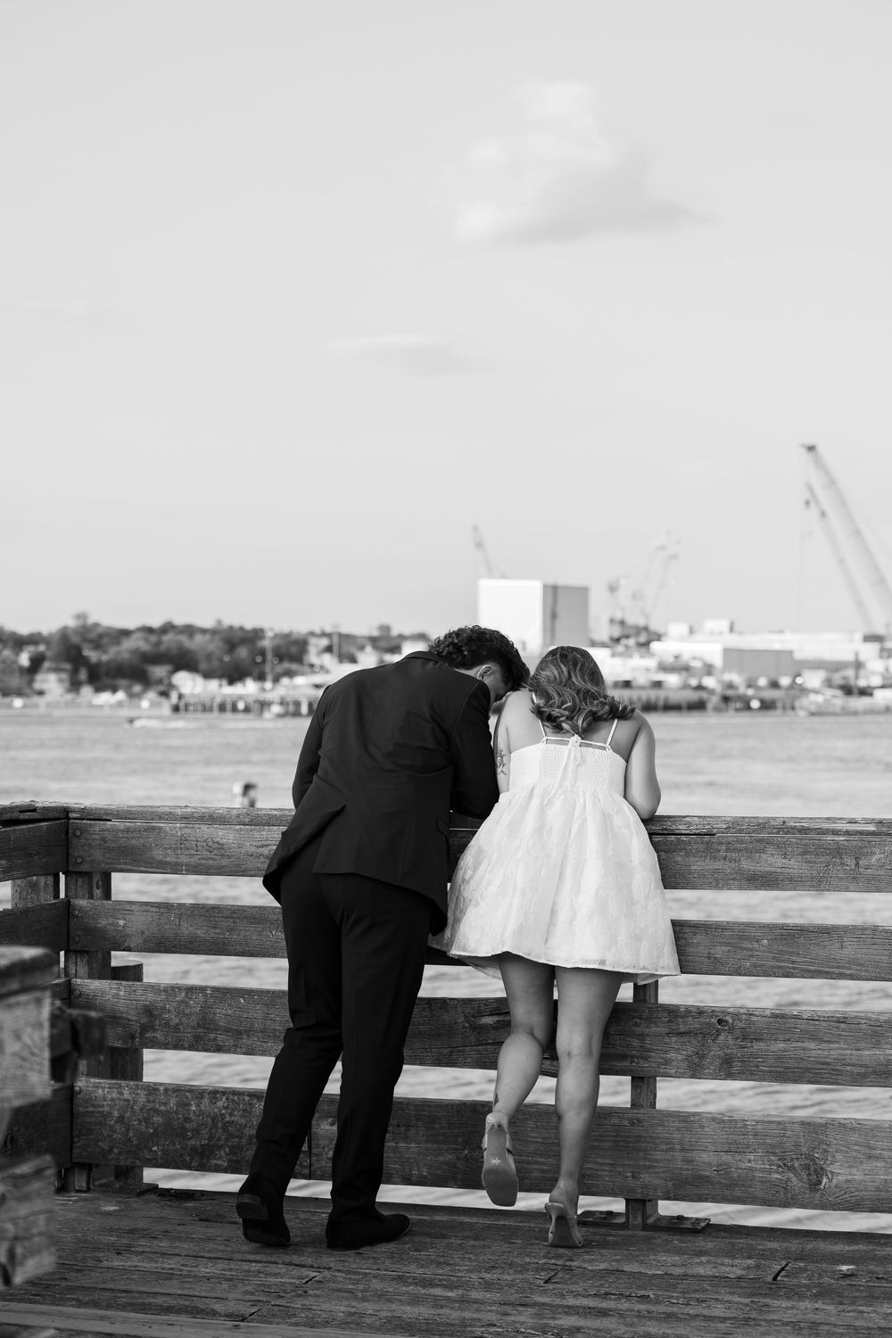 Black and white photo of a couple leaning on a pier railing overlooking the water, man in suit and woman in white dress.