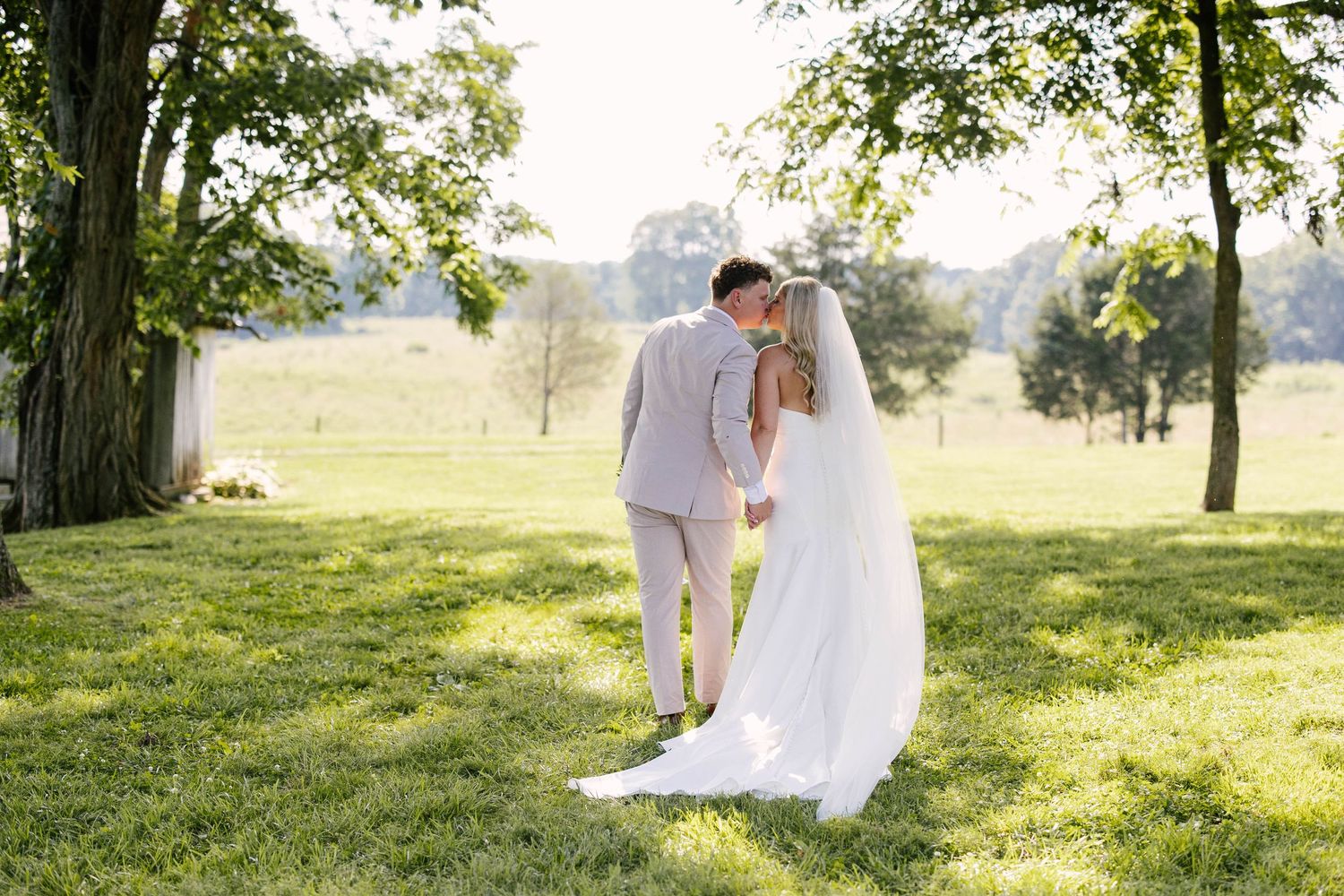 Romantic couple in white attire embraces on a sunlit grassy hillside beneath towering trees during golden hour.