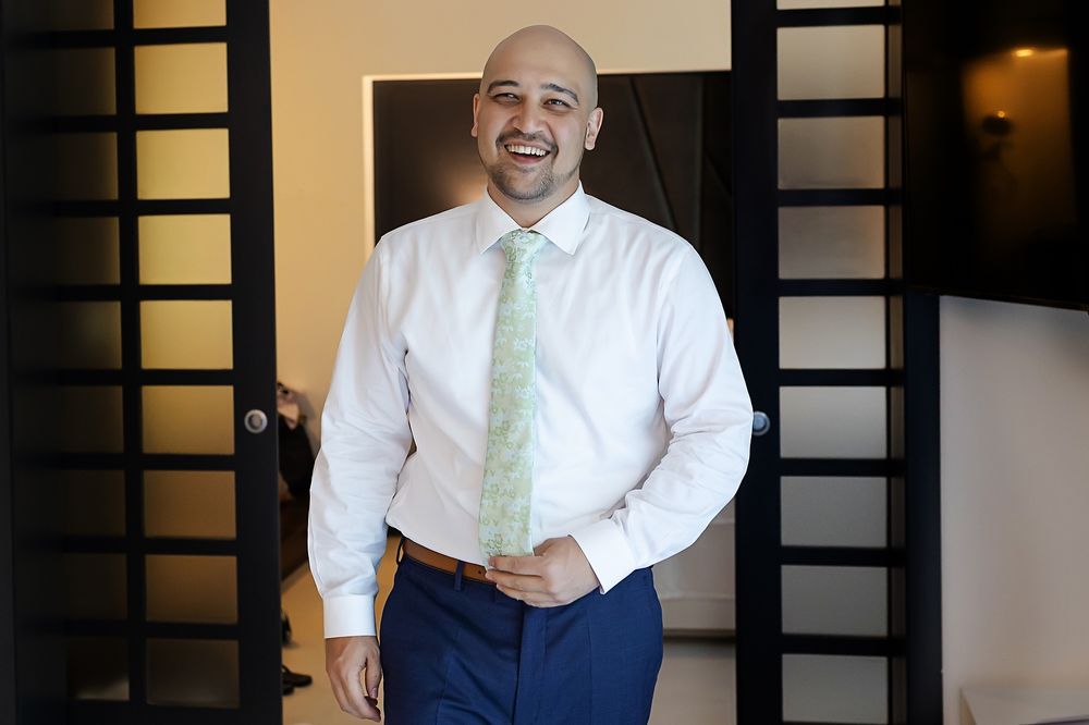 Professional wearing a crisp white dress shirt with mint green tie and navy slacks stands smiling in an office hallway.