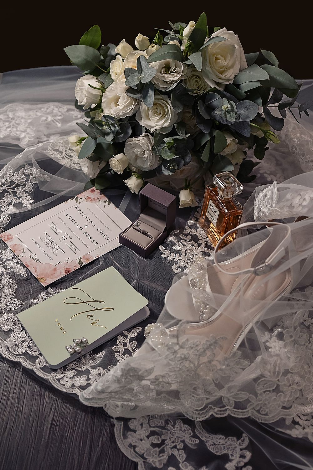 Wedding details including white roses, lace veil, perfume bottles and invitation cards arranged on dark wooden surface.