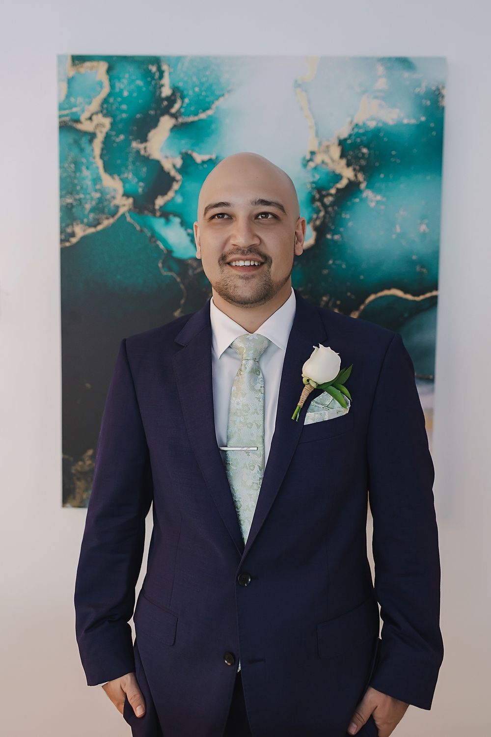 A person in a navy suit and mint green tie poses against a turquoise and gold abstract wall art backdrop.