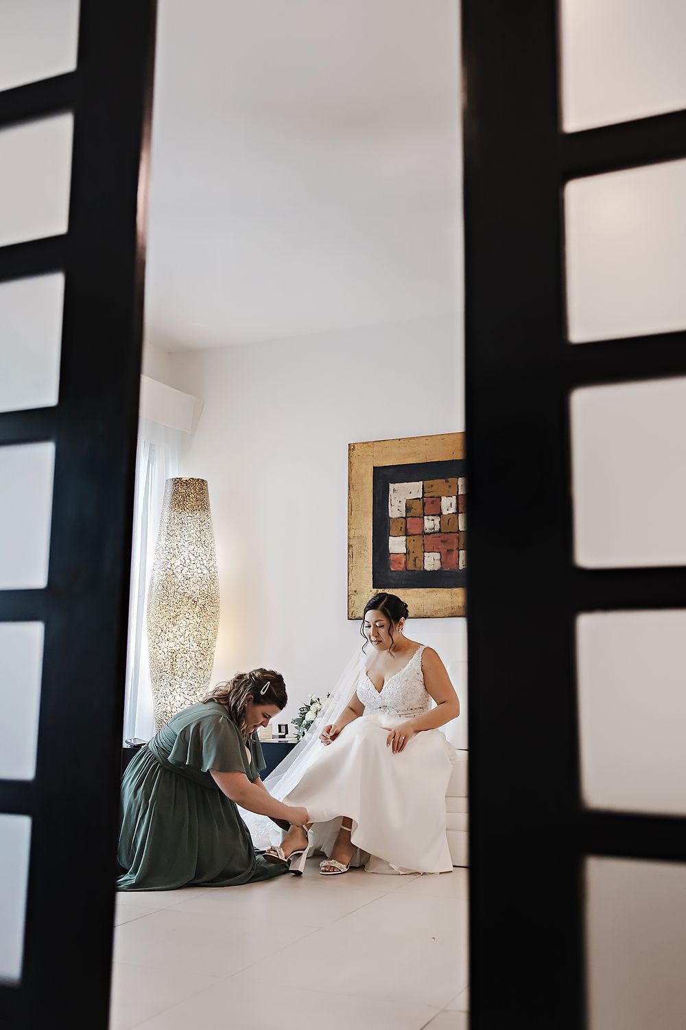 Through dark vertical bars, a figure in white sits on the floor in a minimalist room with pendant lighting and wall art.