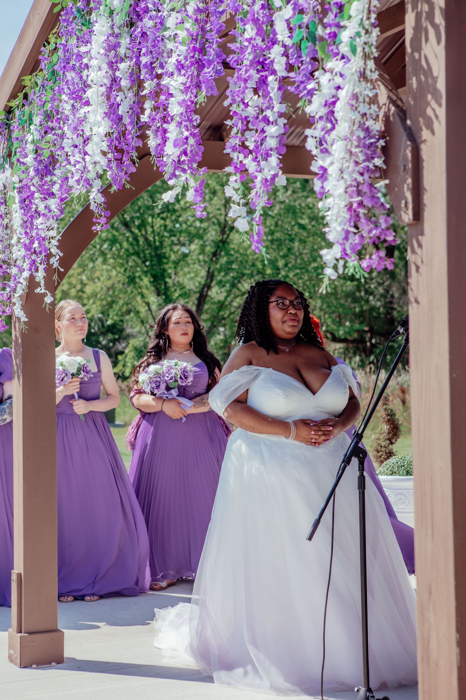 Wedding party poses under beautiful hanging purple wisteria flowers at an outdoor venue.