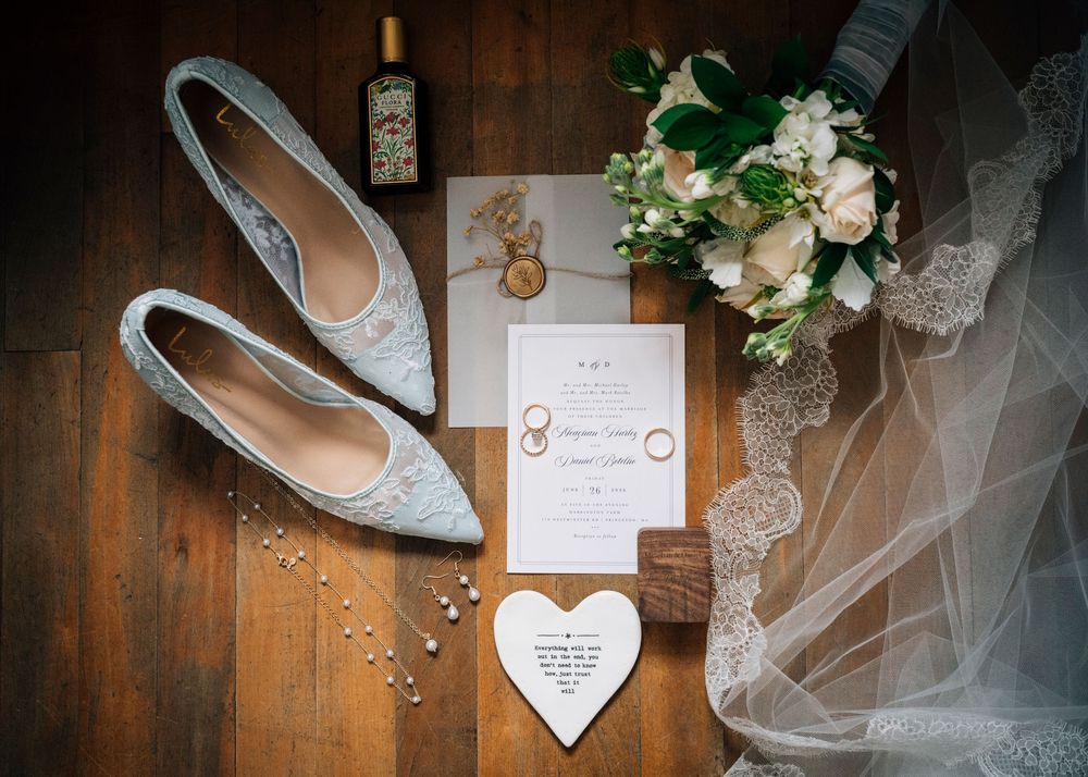 Elegant wedding flat lay with lace heels, floral bouquet, invitation suite, rings, and bridal veil on wood floor.