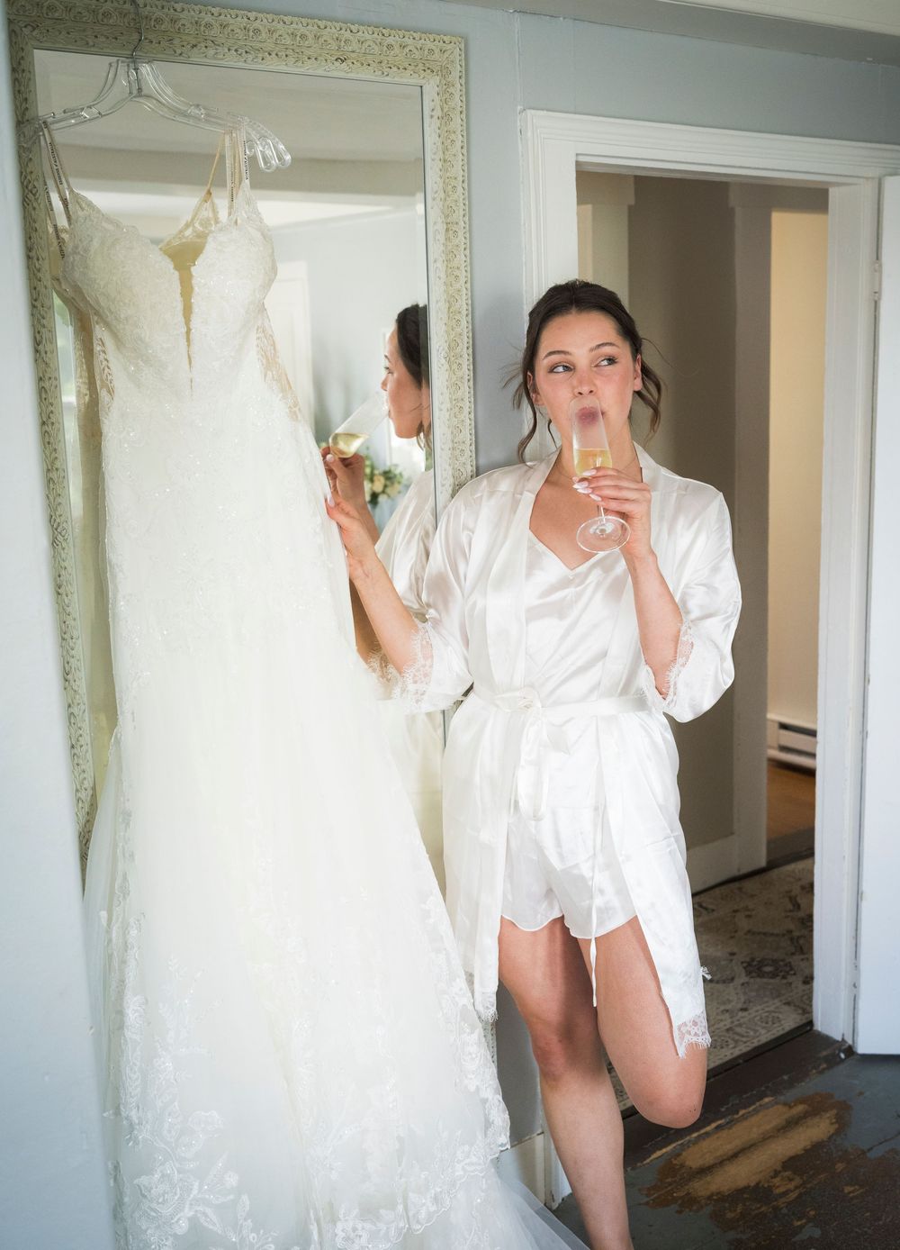 Bride in white satin robe sipping champagne beside her lace wedding gown hanging near a gold-framed mirror.