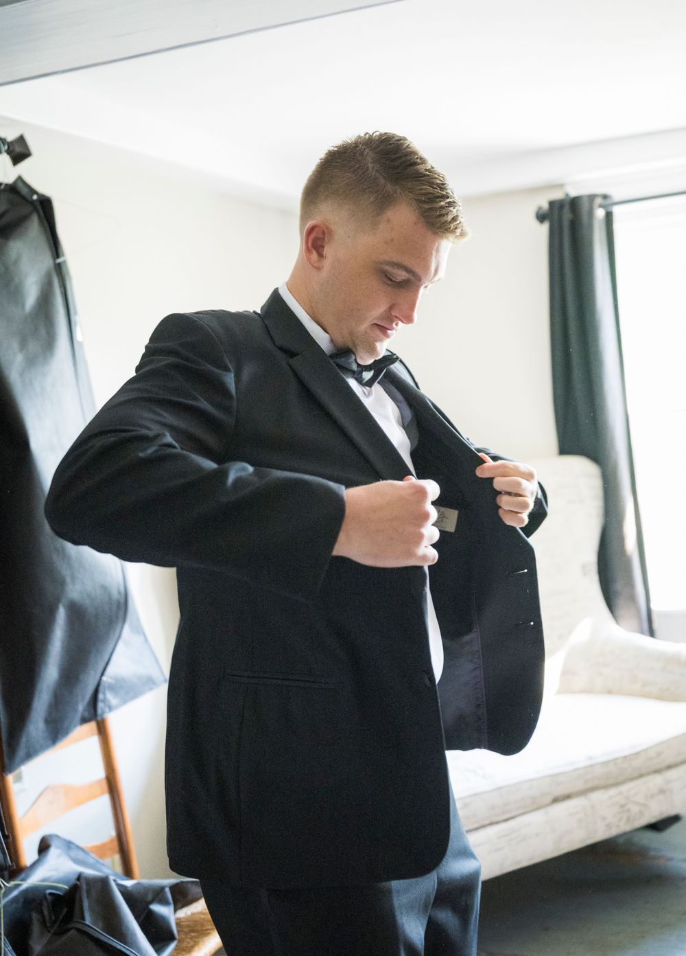 A groom in a black tuxedo and bow tie adjusts his jacket while getting ready indoors.
