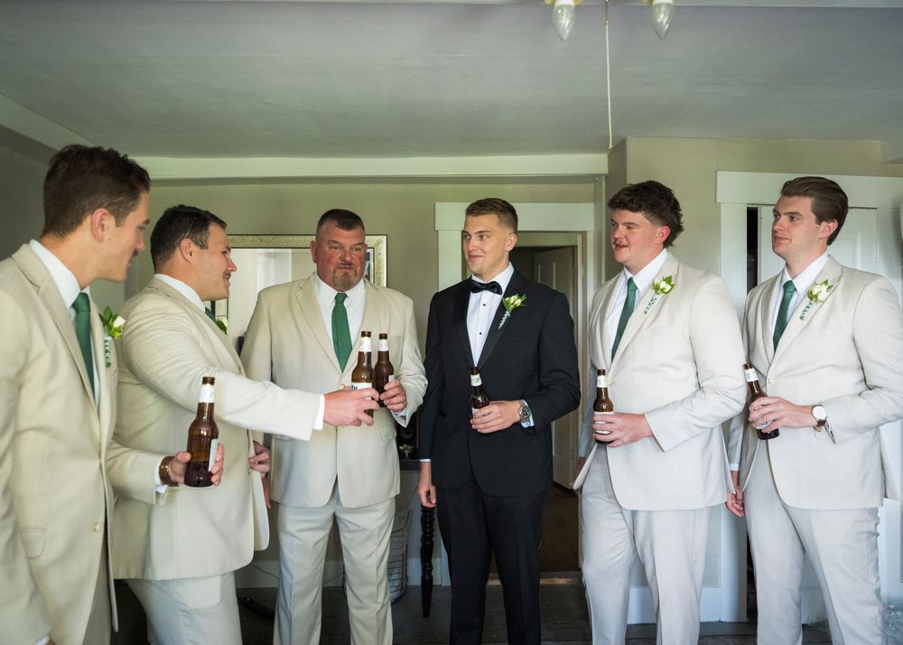 Groom in tuxedo and groomsmen in tan suits share drinks while preparing for wedding ceremony.