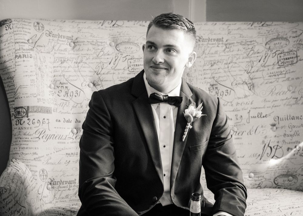Groom in black tuxedo with boutonniere smiling while seated on ornate script-patterned sofa, black and white photo.
