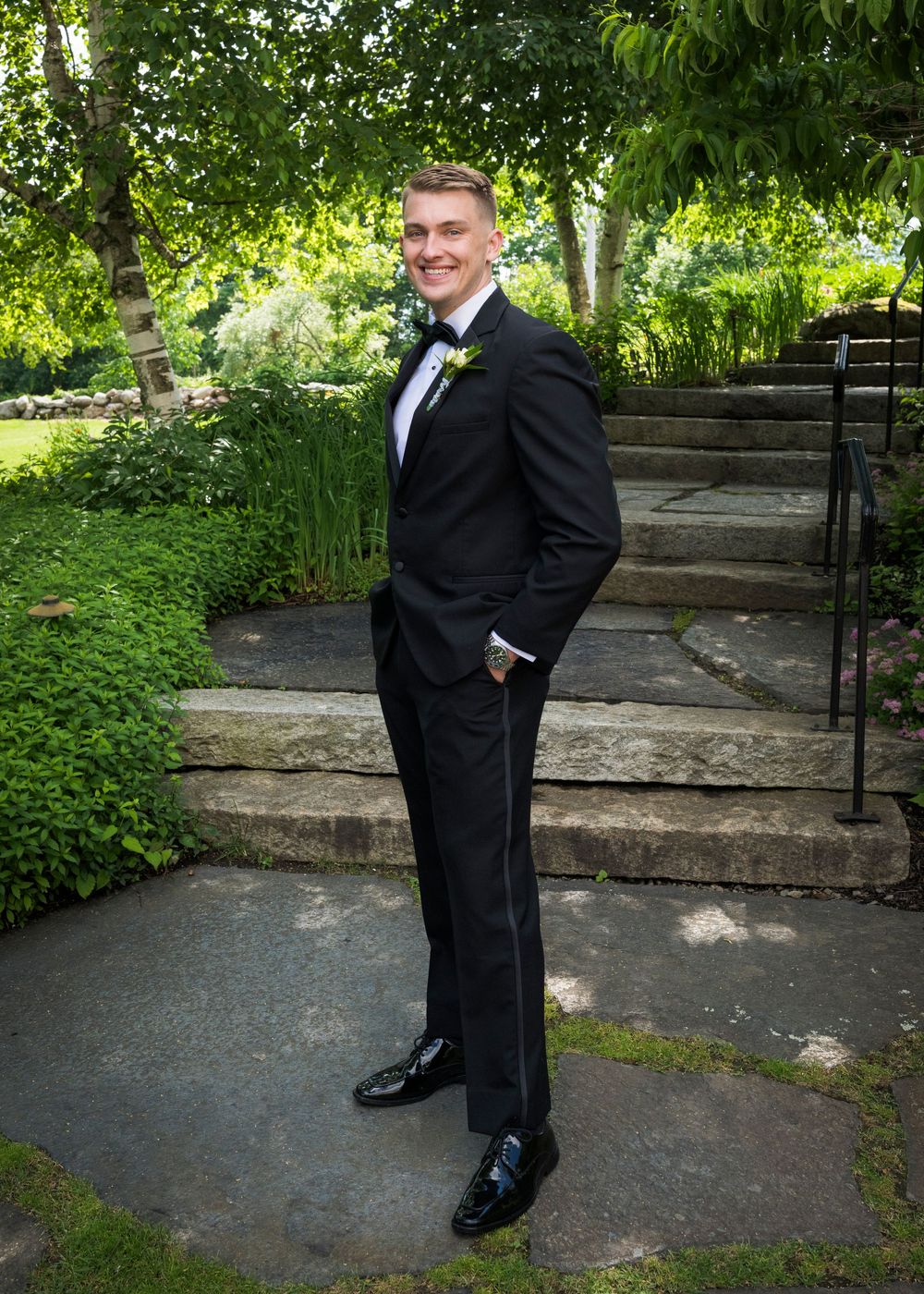 Handsome groom in a black tuxedo poses confidently in a lush garden with stone steps on his wedding day.