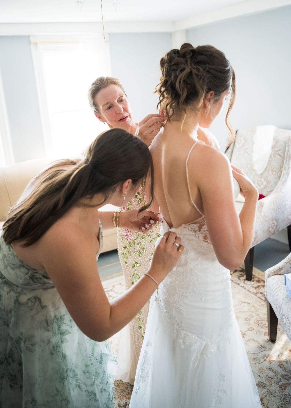Two women helping bride into lace backless wedding gown in a bright bridal suite before the ceremony.