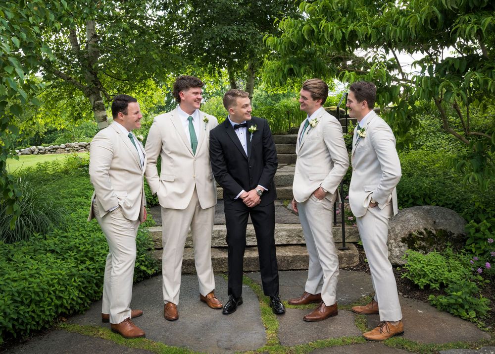 Groom in black tuxedo laughs with four groomsmen in cream suits in lush garden setting.
