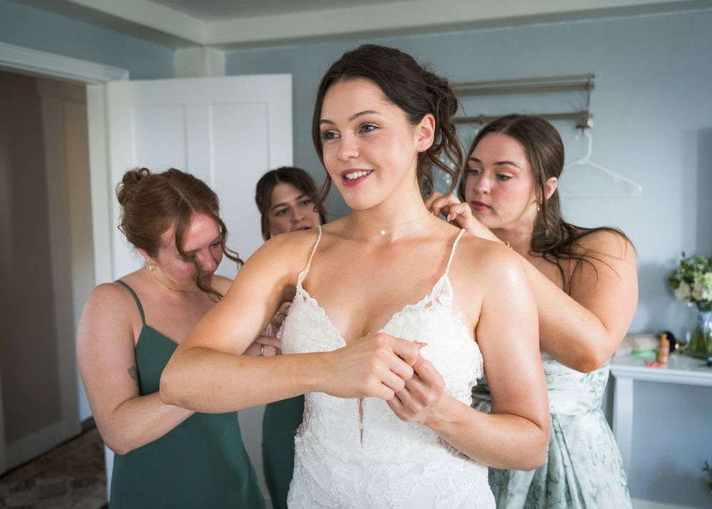 Bridesmaids help the smiling bride into her white lace wedding dress during bridal preparations indoors.