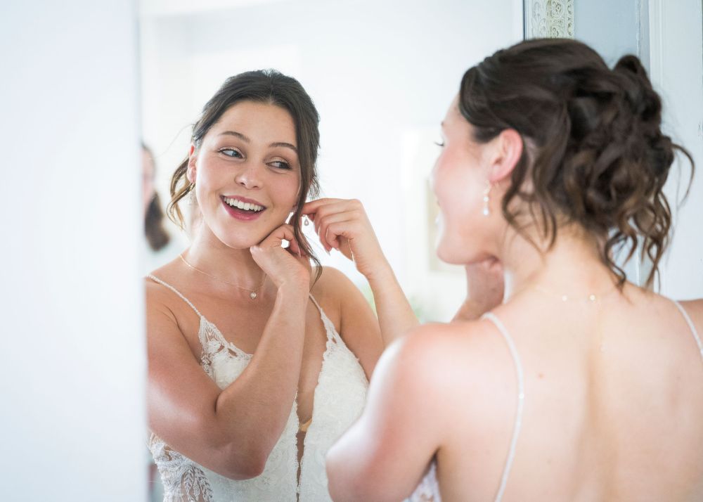 Smiling bride in lace wedding dress putting on earrings while looking at her reflection in a mirror.