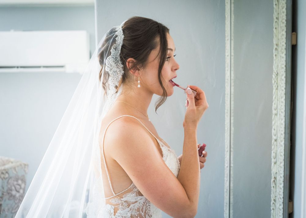 Bride in a lace wedding dress applying lipstick in a mirror while wearing a veil.