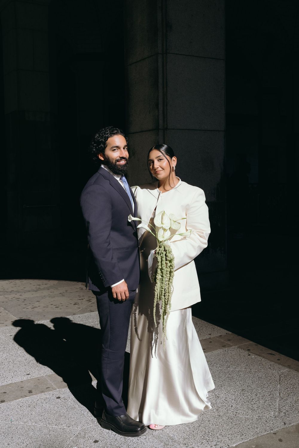 Couple posing in wedding attire, groom in navy suit, bride in white jacket and satin skirt holding floral bouquet.