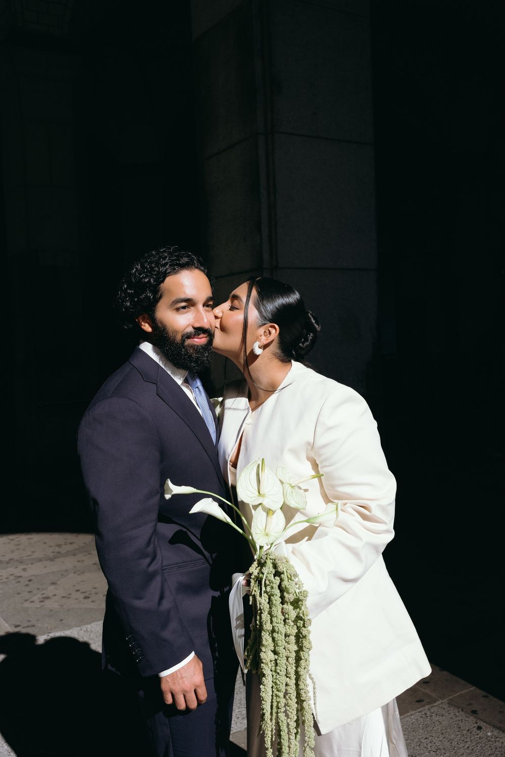 Bride kisses groom on cheek outdoors, she wears white blazer holding calla lily bouquet, he wears navy suit.