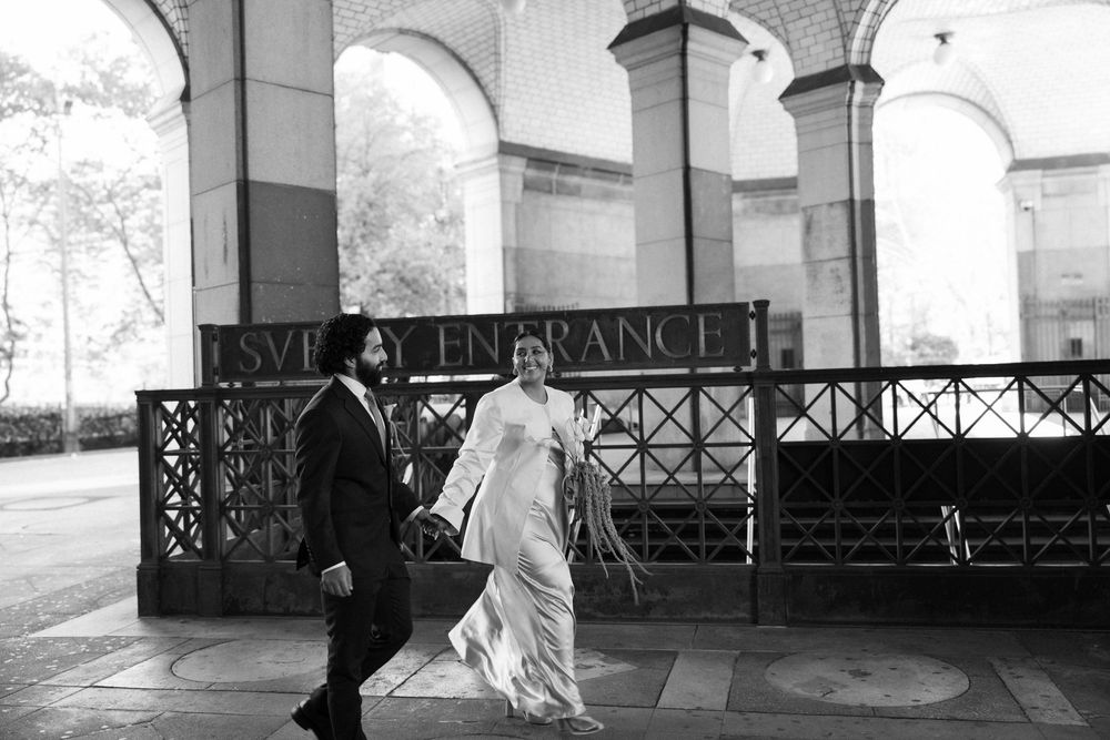 Black and white photo of a couple in wedding attire walking past a building entrance with arches.