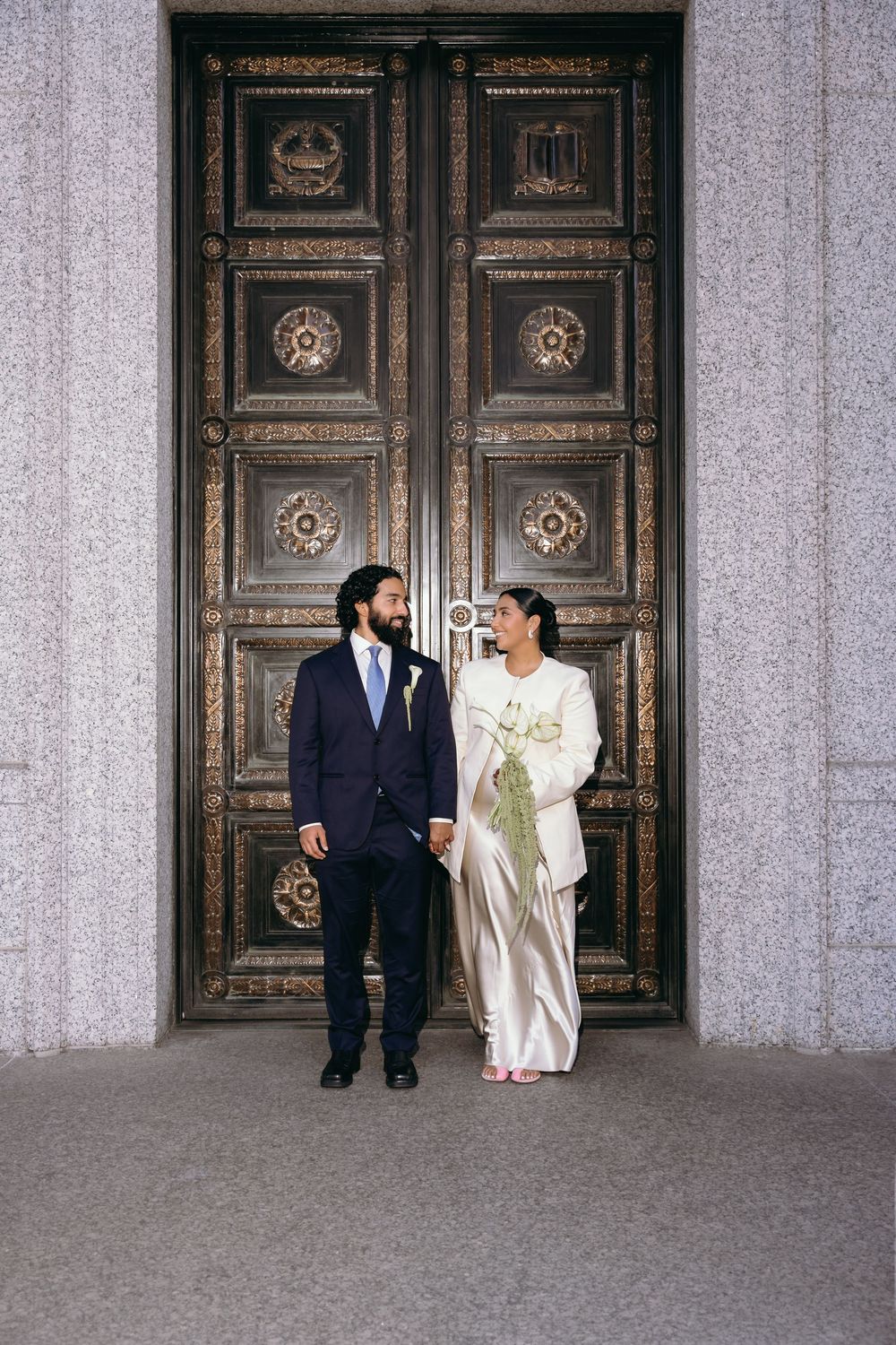 Bride and groom stand holding hands before ornate bronze doors in wedding attire, she in ivory gown, he in navy suit.
