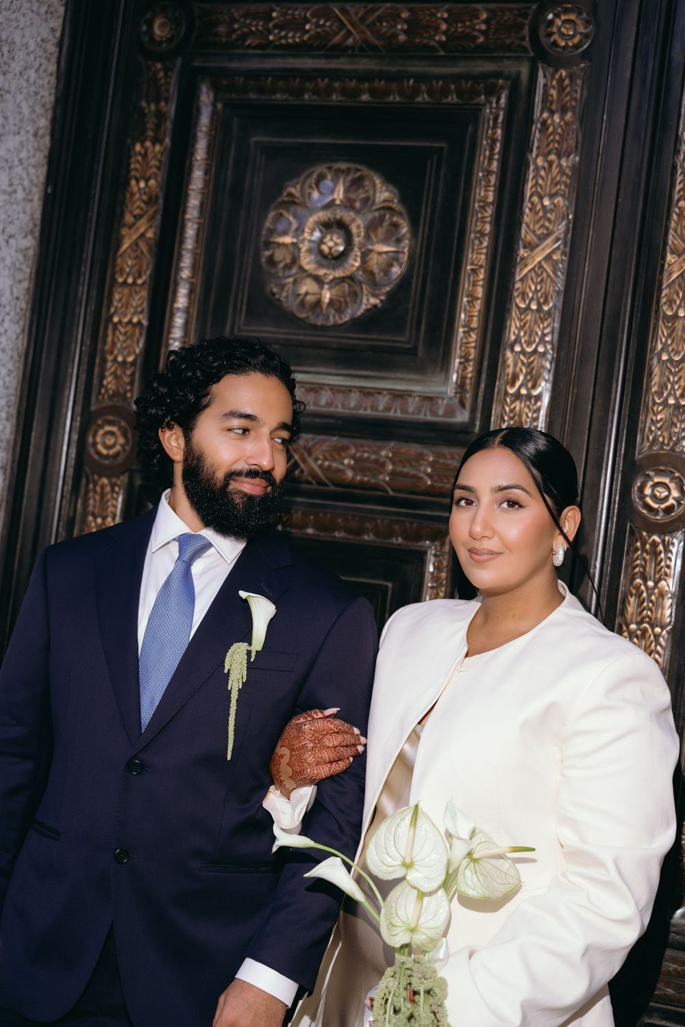 A bearded groom in a navy suit and bride in a white blazer pose elegantly before an ornate carved wooden backdrop.
