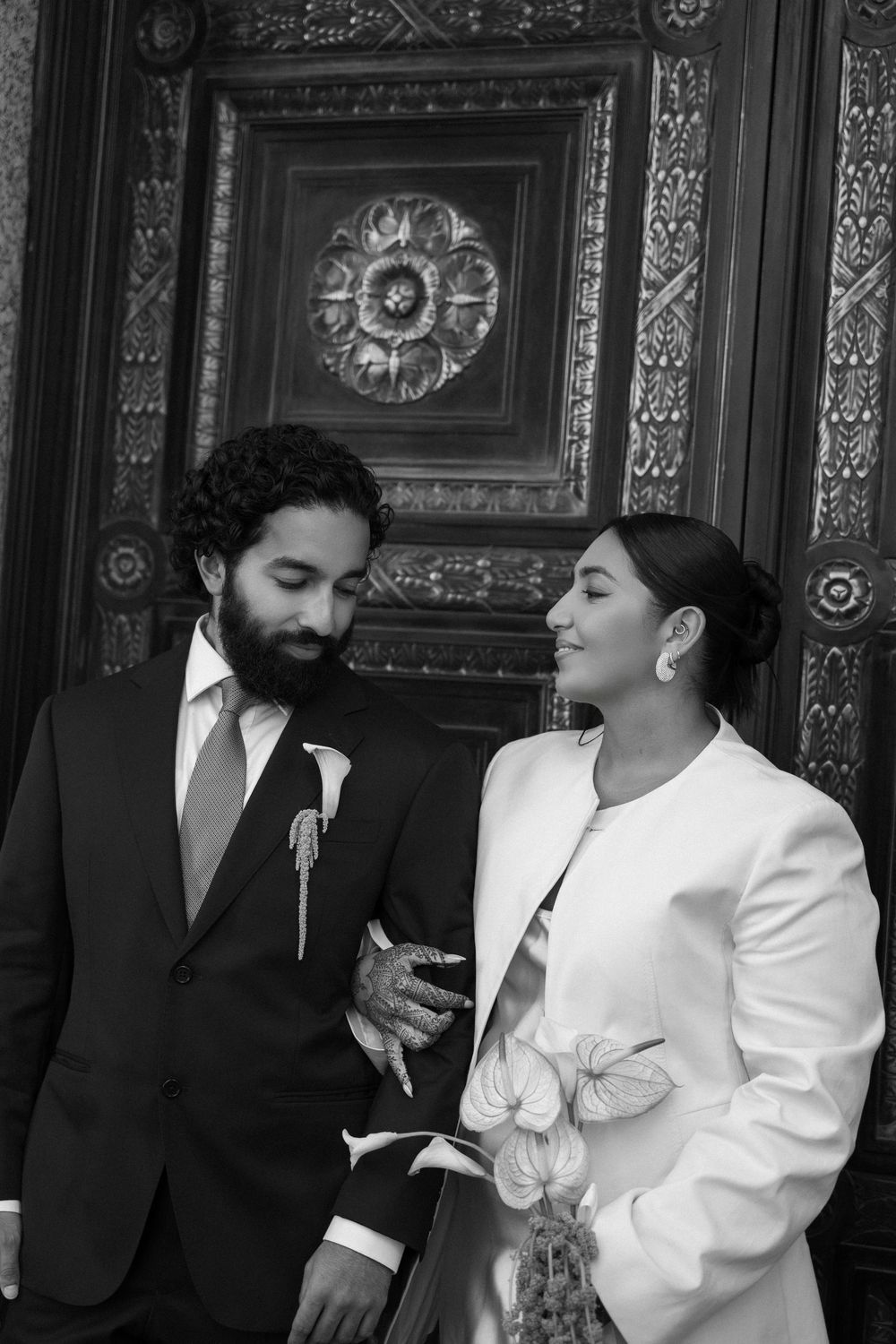 Black and white photo of a couple in wedding attire smiling at each other before an ornate carved wooden door.