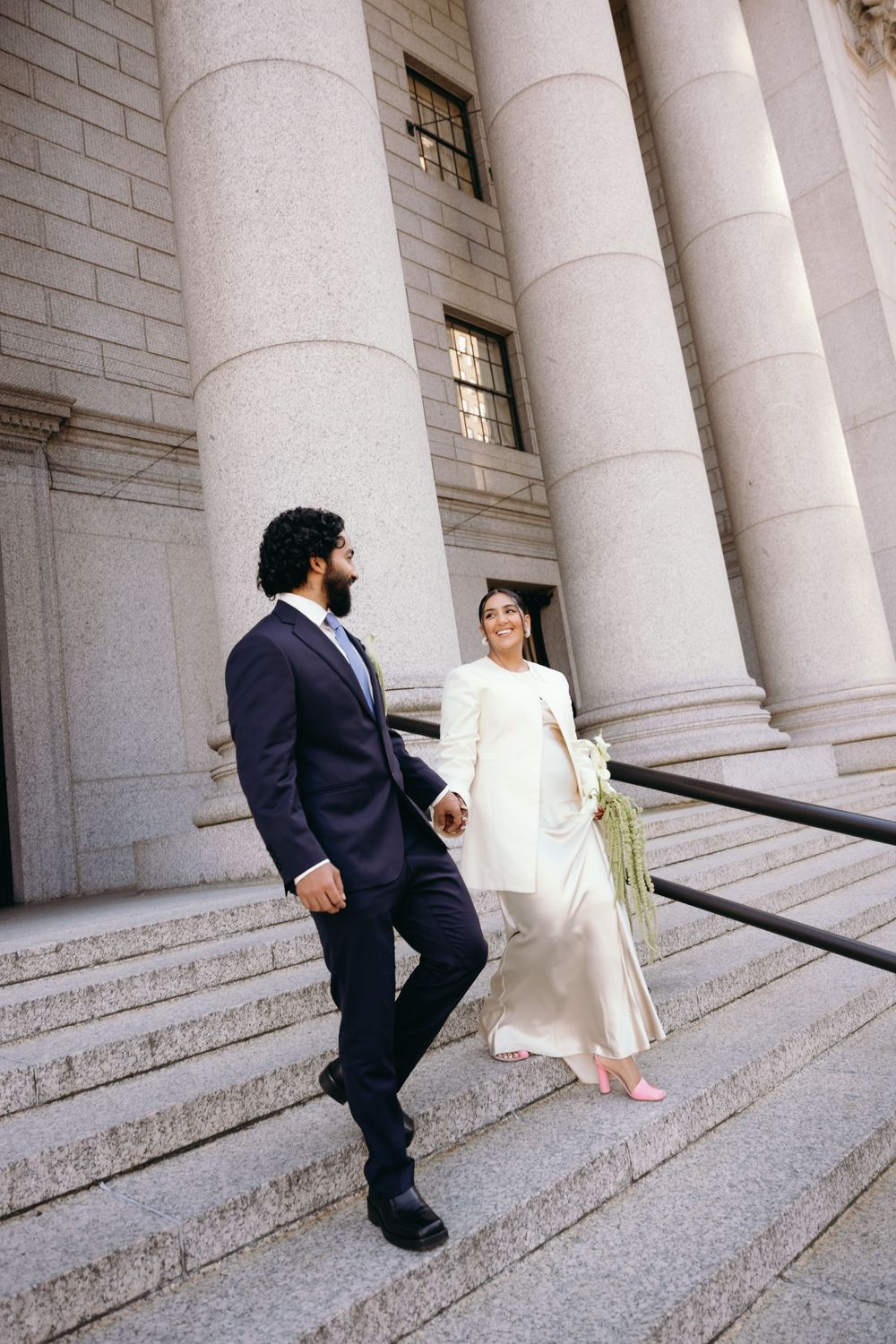 Bride and groom walk down stone steps holding hands beside grand columns in wedding attire.