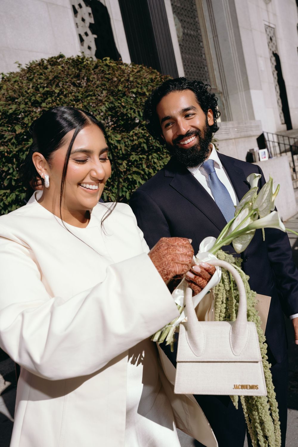 Smiling South Asian bride in white dress holding Jacquemus bag with groom in navy suit behind her.