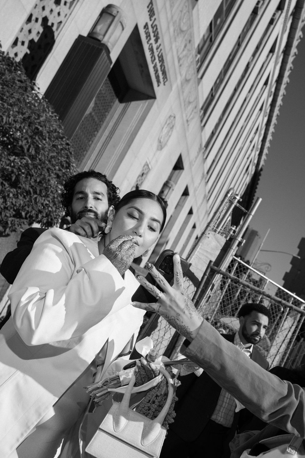 Black and white street photo of a woman in white gesturing with her hand, with people and a building in the background.