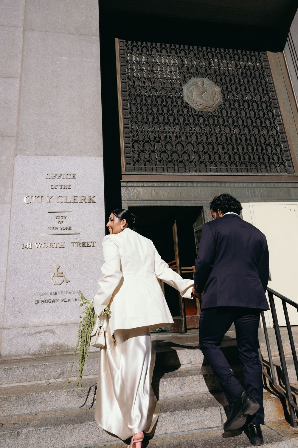 Bride and groom hold hands climbing steps to NYC Office of the City Clerk for their courthouse wedding.