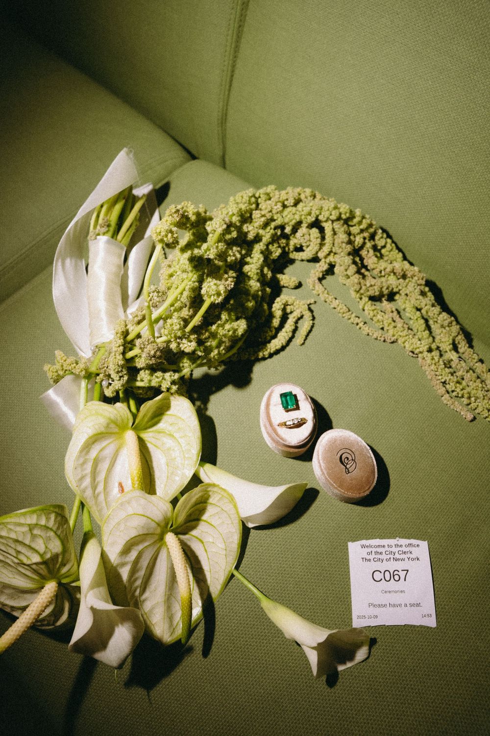 Wedding flat lay with white anthurium flowers, green amaranthus, and an emerald ring on a green velvet chair.