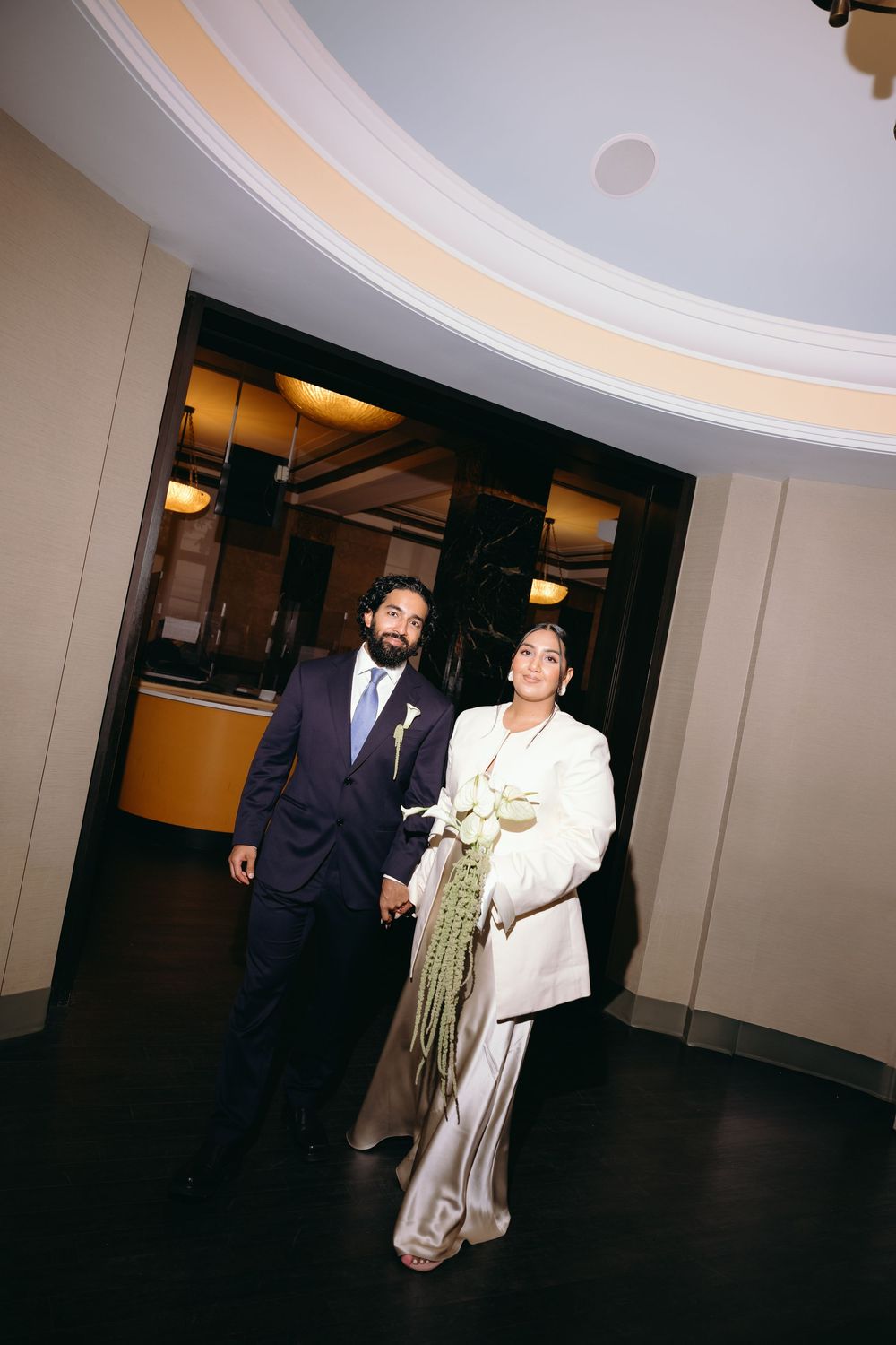 Elegant couple in wedding attire holding hands, bride in white suit with floral bouquet, groom in navy suit.