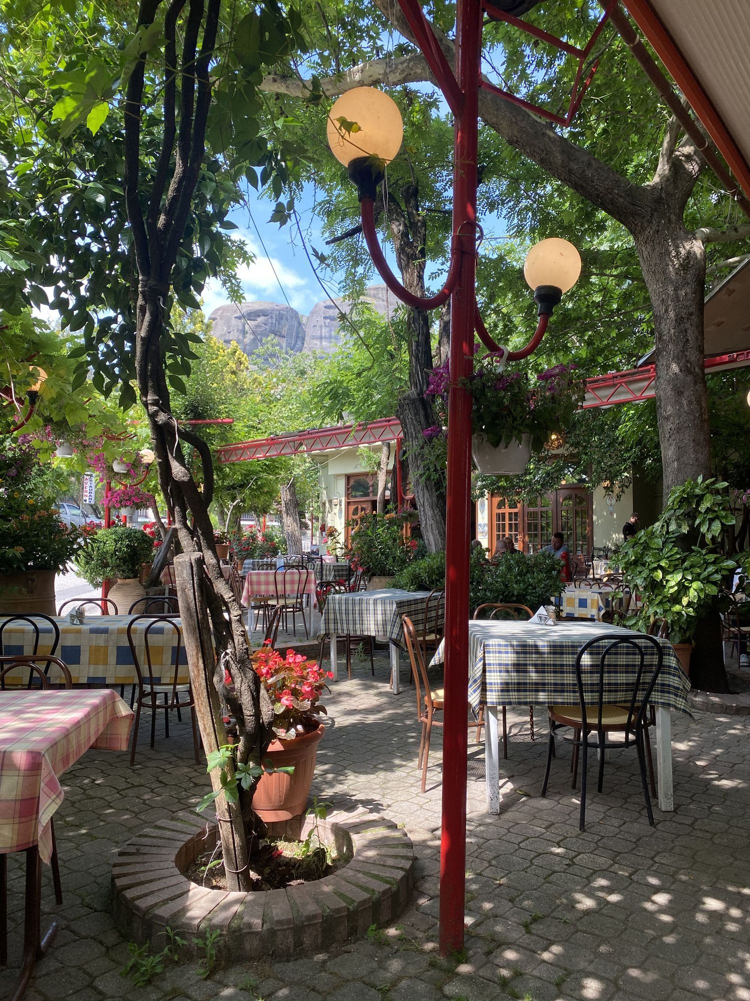 Charming outdoor restaurant patio with red columns, hanging lanterns, and shaded seating areas under lush green trees.
