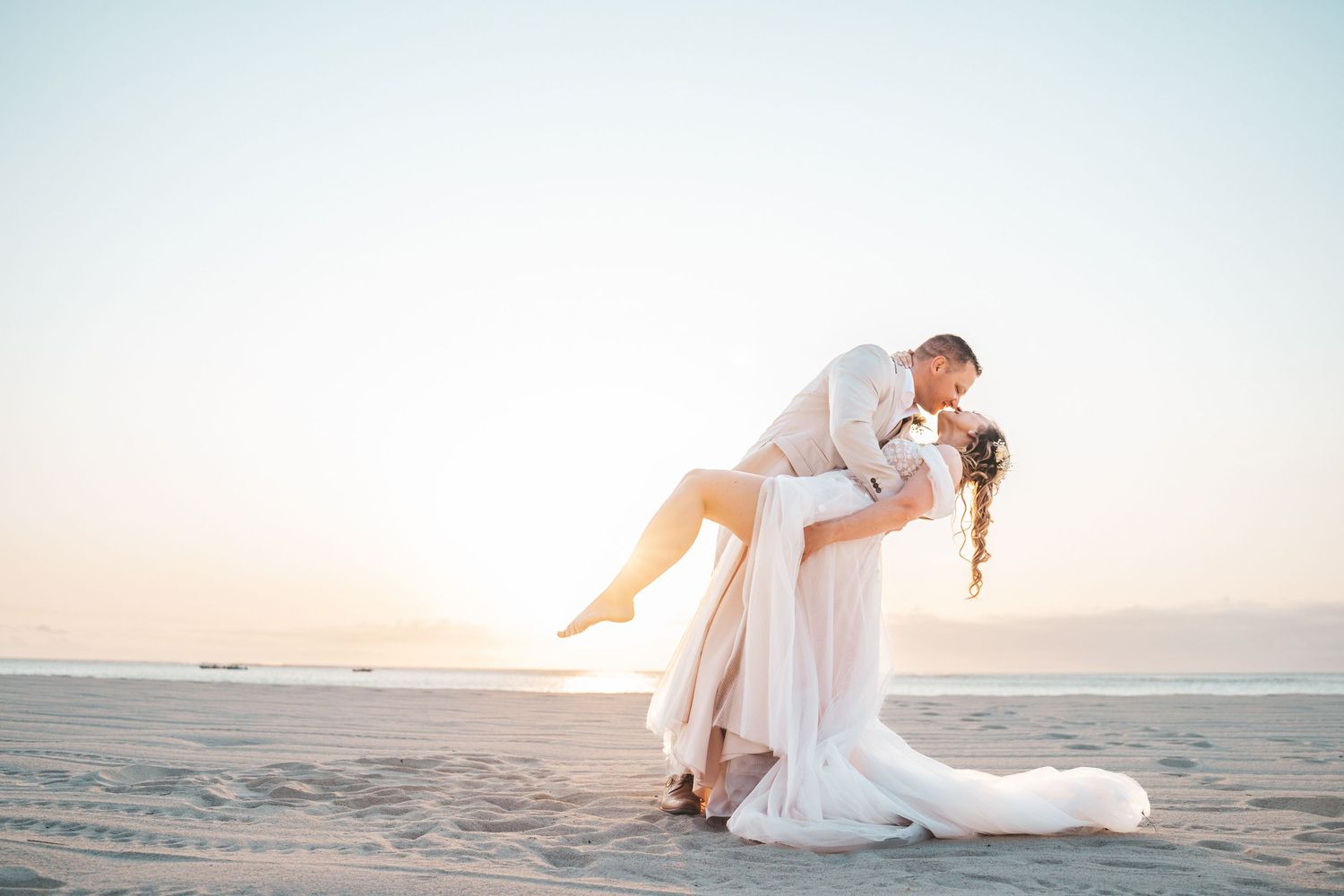 Romantic beach wedding photo of couple embracing during sunset with flowing white dress.