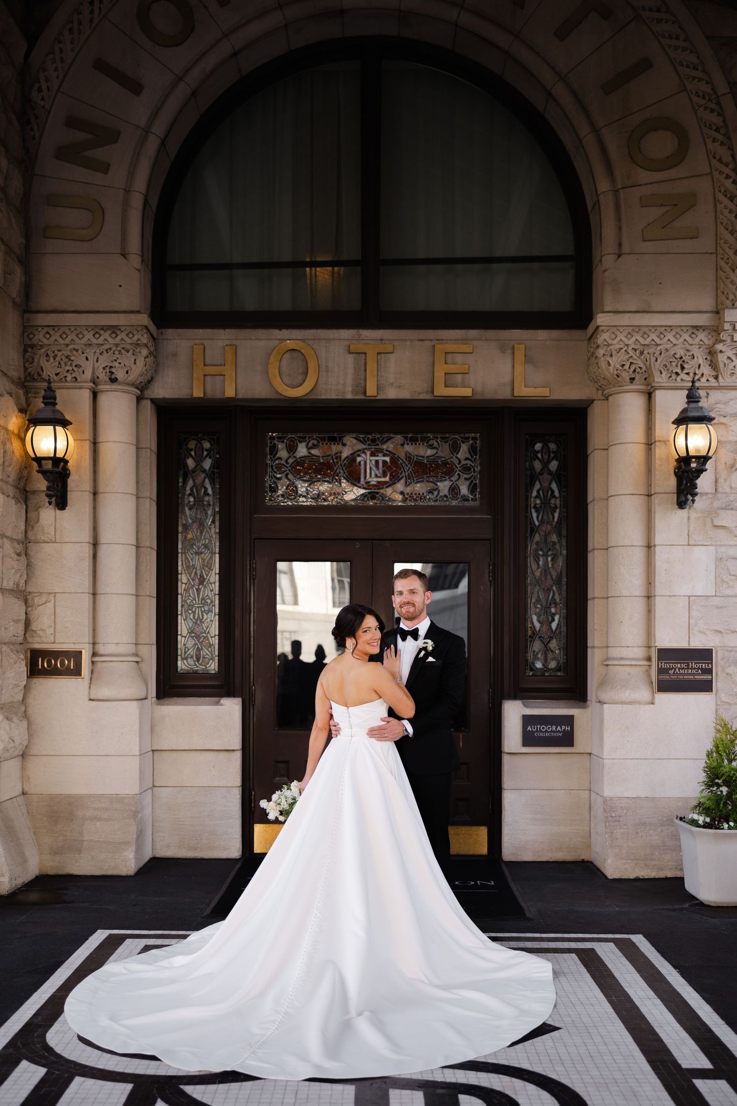 A couple poses in front of an elegant hotel entrance with ornate stone architecture and glowing wall sconces.