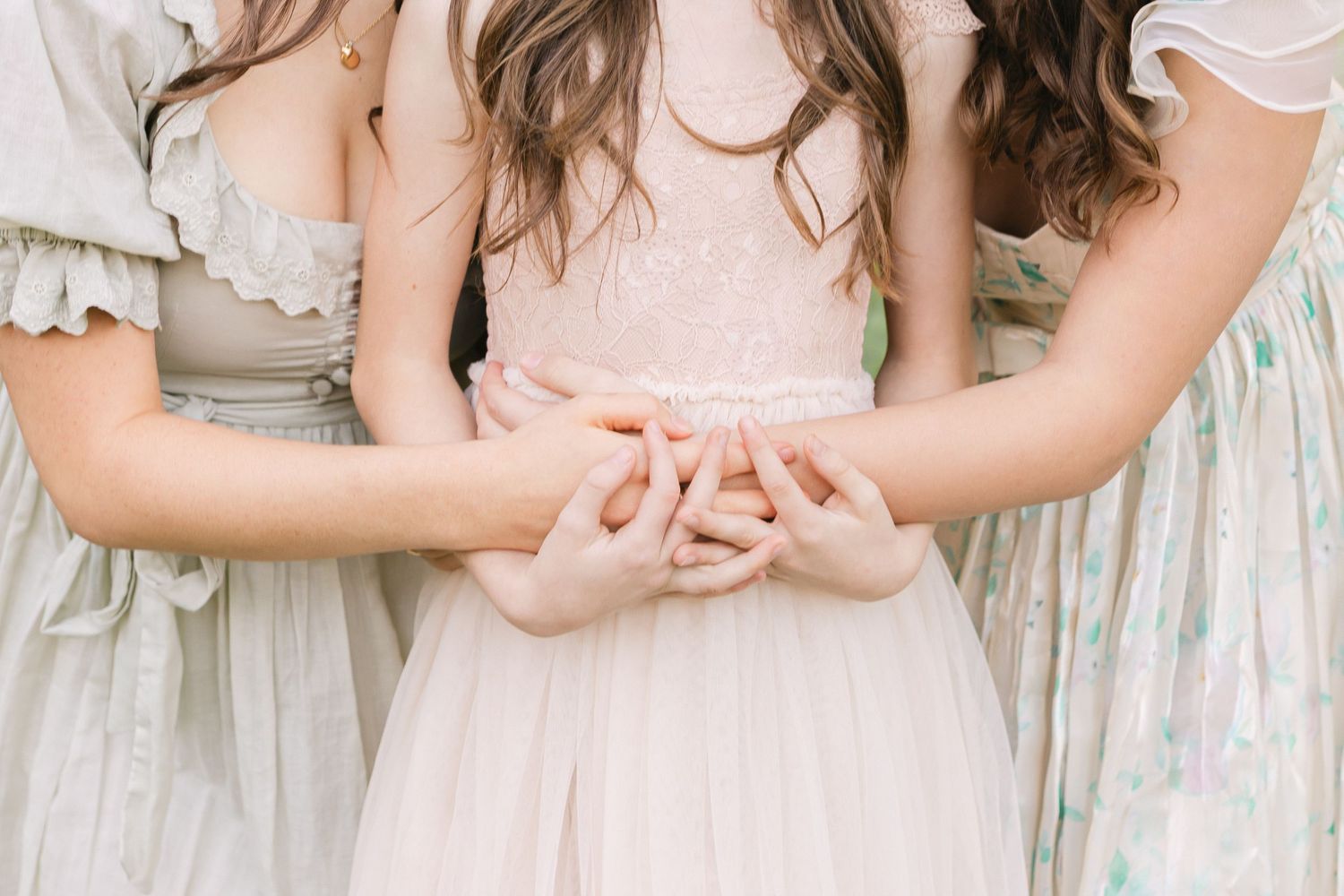 Three people wearing pastel vintage-style dresses hold hands together in a delicate moment.