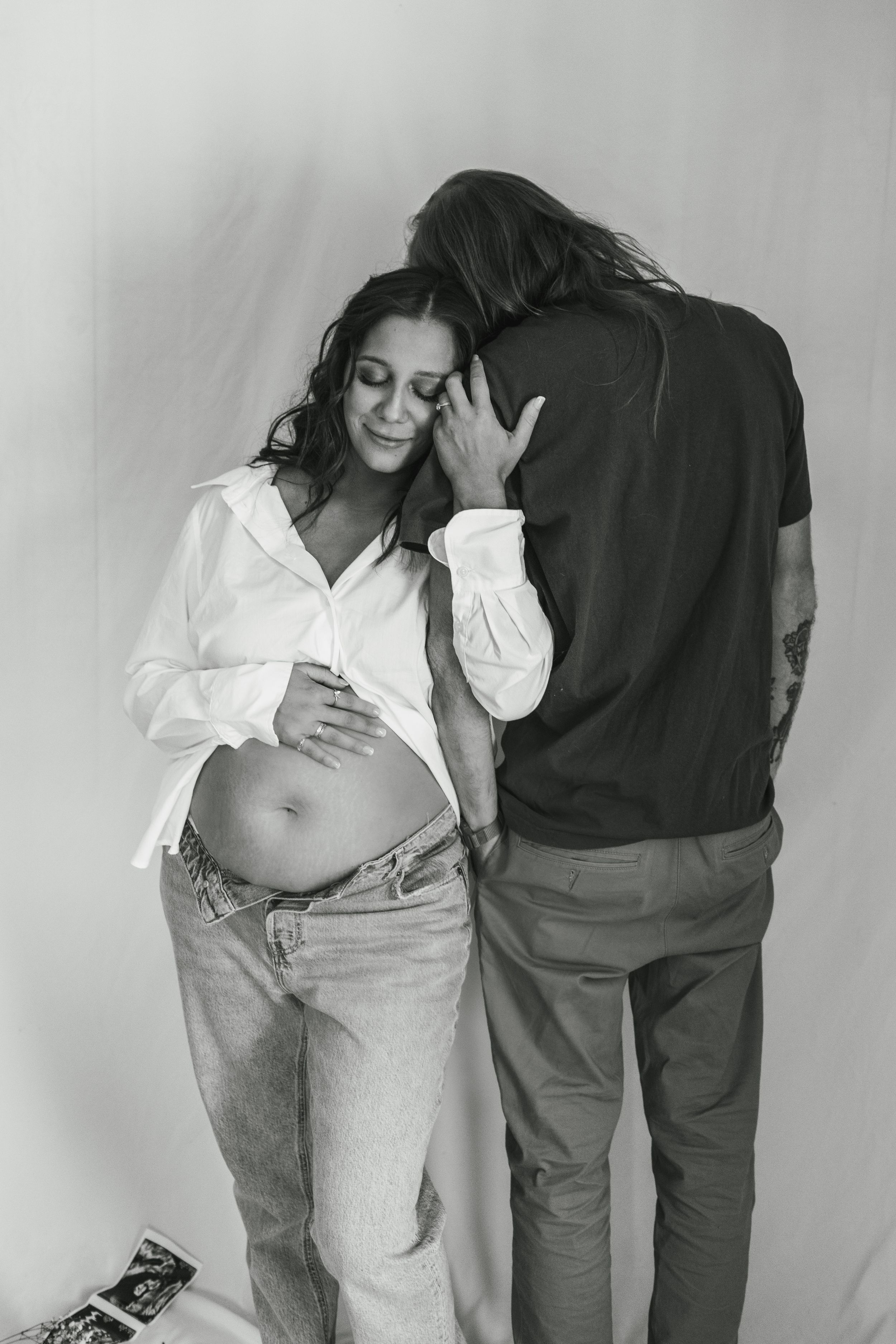 Intimate maternity portrait showing expectant couple in casual attire against white background.