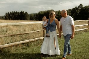 A young family with a baby walks along a wooden fence in a green field on a cloudy day.