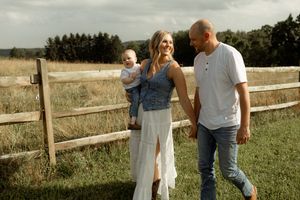 Young family with baby poses by rustic wooden fence in green meadow during outdoor countryside photo session.