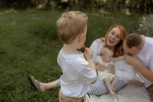 A young boy offers a flower to his laughing redhead mother and father sitting on a blanket in a lush green park.