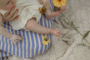 Baby in white top and blue striped pants holding yellow flowers on a white knit blanket.