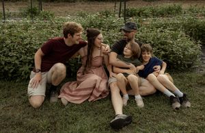 A happy family of five sitting together on green grass outdoors, smiling and embracing in a casual photo.