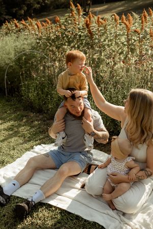 Family enjoying a sunny outdoor picnic on a blanket with two young children among tall wildflowers in a park.