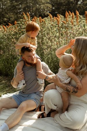 A young family enjoys a sunny outdoor picnic, with dad holding a toddler on his shoulders and mom cradling a baby.