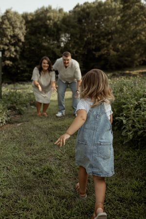 A young girl in denim overalls runs toward her smiling parents on a lush green garden path.