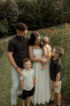 A happy family of five poses outdoors in a lush green field, with parents and three young children dressed in coordinated outfits.