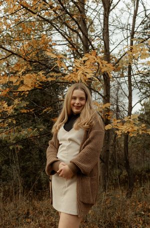 Young blonde woman smiling in autumn forest wearing cream dress and brown knit cardigan among golden fall foliage.