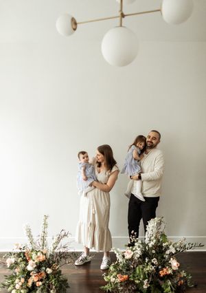 A family in white and dark clothing poses together in a studio with floral arrangements on the floor.