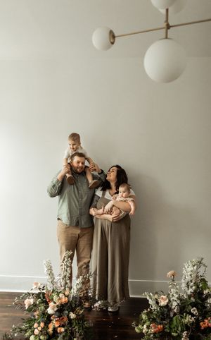 A family poses together in a minimalist studio setting with floral arrangements and white globe lights.