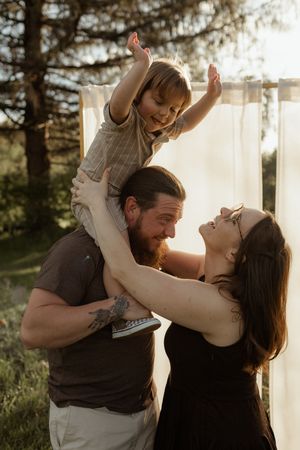 A joyful family outdoors at golden hour, father lifting toddler while mother laughs, with soft white curtains and trees behind.
