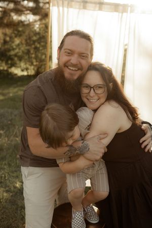 A bearded man and woman with glasses share a warm family hug with a young child outdoors in golden sunlight.